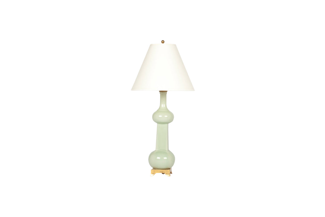 A single small Hadley lamp in blue celadon glaze with a 23k gold base, brass harp and dimmer socket, and off white vellum paper shade, on a white background.