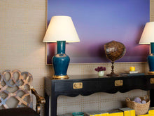 A pair of Garniture table lamps in Prussian Blue glaze with 23k gold bases, and off white vellum paper shades, flanking a side table in a living room.